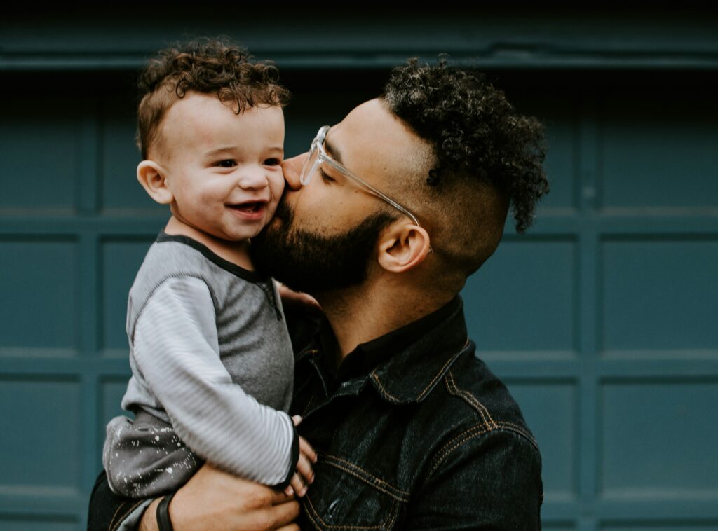 Father kissing his smiling young child while holding them in his arms – symbolising the emotional depth and challenges of modern fatherhood