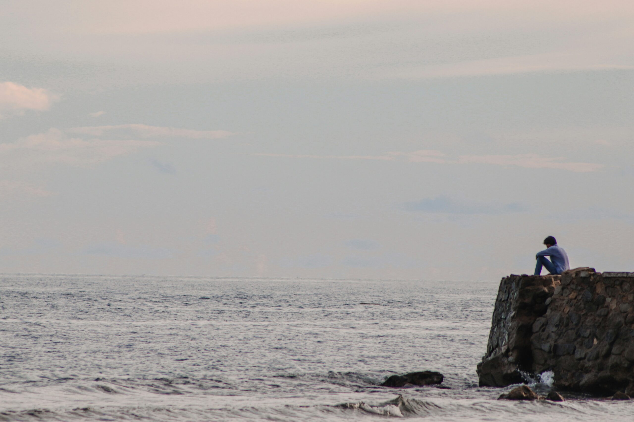 Man sitting alone on a rocky ledge by the sea, gazing out over the water – symbolising isolation, reflection and the hidden experience of depression in men