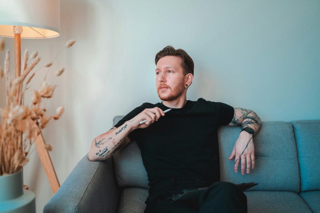 Thoughtful man with tattoos sitting on a sofa holding a stylus – representing modern masculinity and the value of choosing a male therapist for relatable support